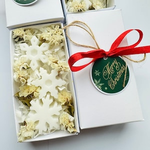 Pode incluir: Velas brancas em forma de floco de neve em uma caixa branca com flores brancas secas. A caixa é amarrada com uma fita vermelha e uma etiqueta verde que diz "Feliz Natal".