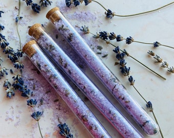 May include: Three glass test tubes filled with lavender bath salts and topped with cork stoppers. The test tubes are arranged in a row on a white surface with dried lavender sprigs scattered around them.