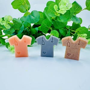 May include: Three scrubs-shaped soap bars in peach, grey, and brown. Each soap has a stethoscope and heart design. The soaps are set against a backdrop of green foliage, creating a natural aesthetic.