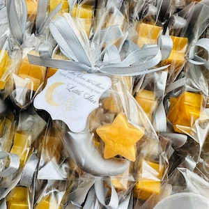 Può includere: Primo piano di bomboniere per baby shower. Ogni bomboniera è un piccolo sapone giallo a forma di stella all'interno di un sacchetto di cellophane trasparente, legato con un nastro argentato. Un'etichetta recita "Over the Moon, Thank you for celebrating our Little Star."