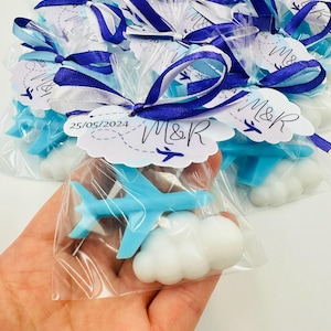 Peut inclure: Un ensemble de savon en forme d'avion bleu clair et de nuage blanc dans un sachet en plastique transparent. Le sachet est noué avec un ruban bleu foncé et blanc et porte une étiquette blanche festonnée imprimée avec 25/05/2024, M&R et un avion.