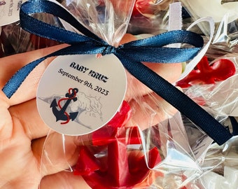 May include: A set of small red and white anchor-shaped favors tied with blue and white ribbon. Each favor has a round tag with the text "Baby Cruz September 9th, 2023" and a small anchor illustration.