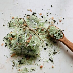 May include: A clear plastic bag filled with green bath salts and tied with twine. The salts are scattered around the bag on a white surface. A wooden spoon with green bath salts is also in the image.