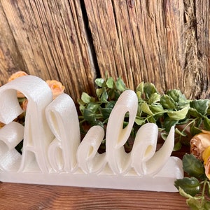 May include: White 3D-printed name sign spelling "Adele" on a wooden surface with a floral arrangement.