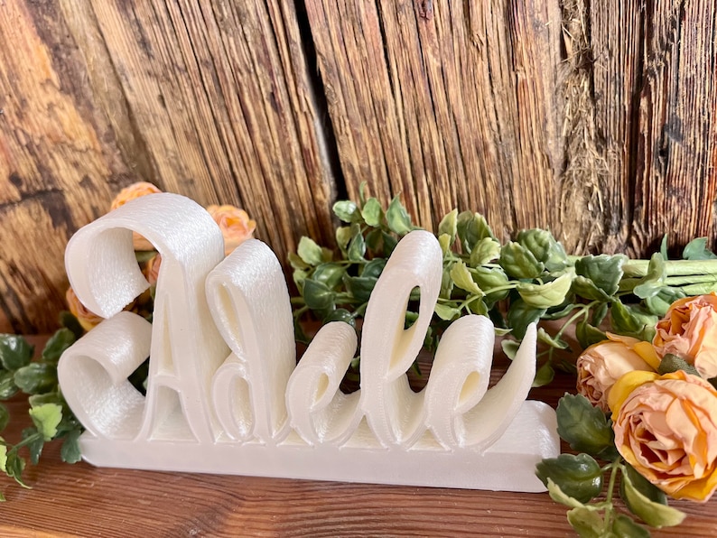 May include: White 3D-printed name sign spelling "Adele" on a wooden surface with a floral arrangement.