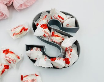 Pode incluir: Um prato de doces em forma de letra S cinza cheio de doces de chocolate branco embrulhados em papéis vermelhos e brancos com a palavra "Raffaello" impressa neles.