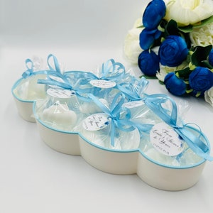 May include: A white cloud-shaped dish filled with small white favors tied with blue ribbon and a tag that says "From Heaven to Yours". The dish is surrounded by a bouquet of white and blue flowers.