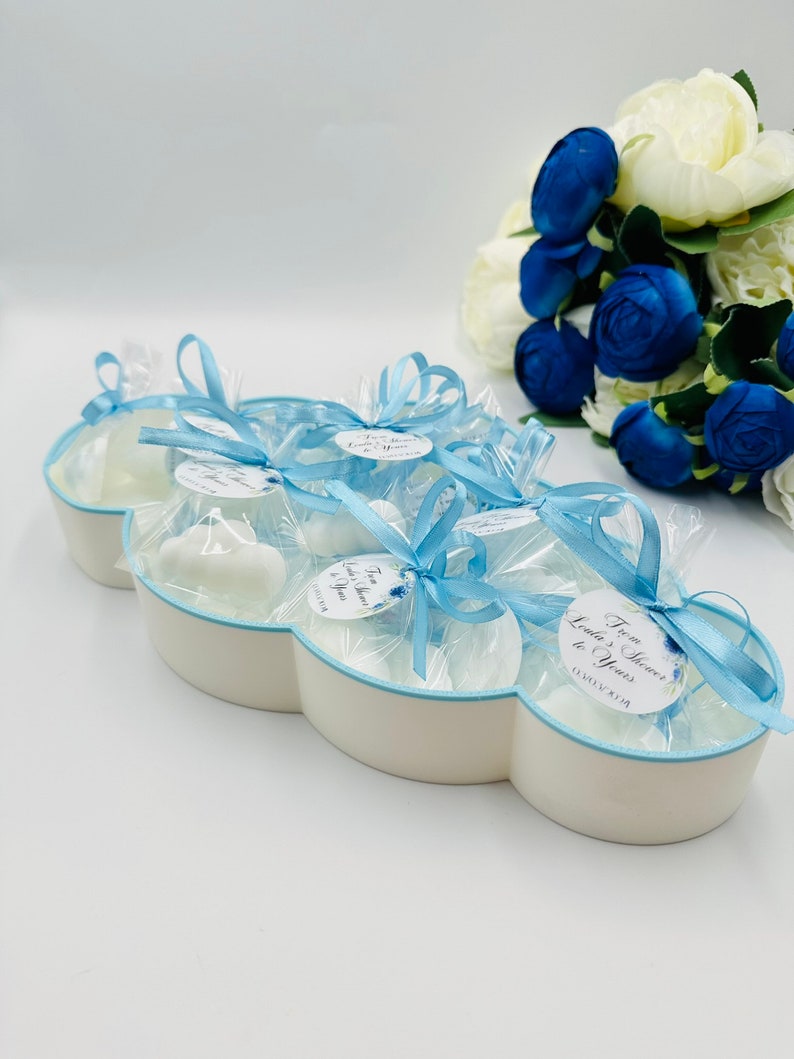 May include: A white cloud-shaped dish filled with small white favors tied with blue ribbon and a tag that says "From Heaven to Yours". The dish is surrounded by a bouquet of white and blue flowers.