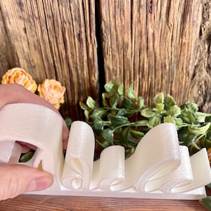 May include: White 3D printed word "Belle" in a cursive font. The word is made of a thick, white material and is sitting on a wooden surface.
