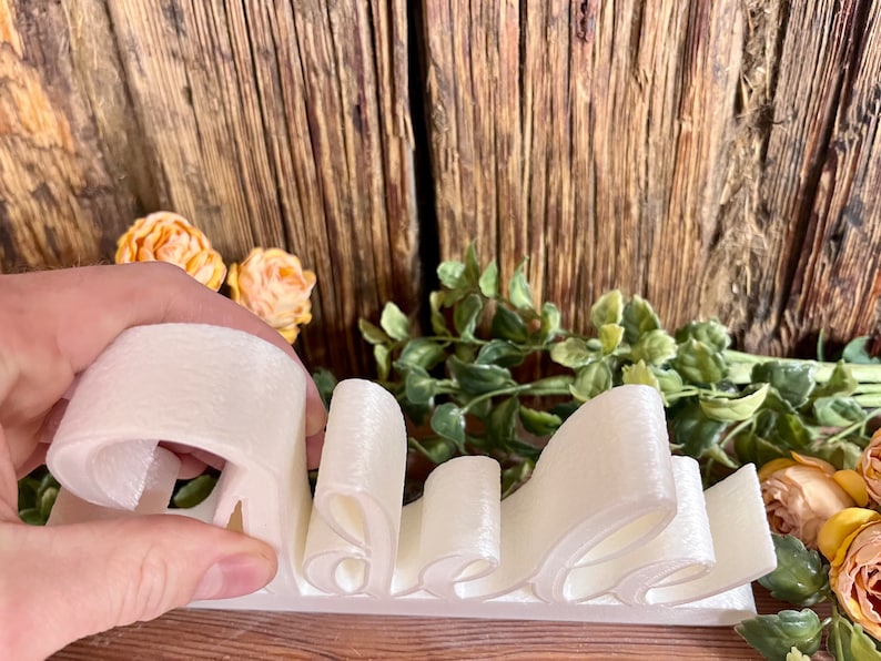 May include: White 3D printed word "Belle" in a cursive font. The word is made of a thick, white material and is sitting on a wooden surface.