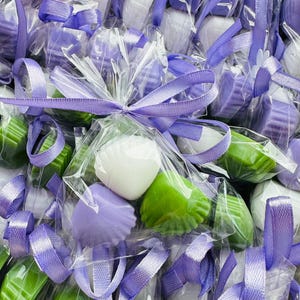 May include: A collection of wrapped soaps in diverse shapes and colours, such as white, green, and lavender. Each soap is individually wrapped in clear cellophane and tied with a lavender ribbon, creating a gift-like presentation.