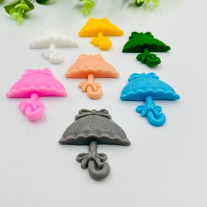 Könnte beinhalten: Sortiment an bunten, miniatürlichen Regenschirm-förmigen Silikon-Anhängern mit Schleifen-Detail. Die Anhänger sind in verschiedenen Farben, darunter rosa, blau, gelb, weiß, grau und grün.
