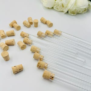 May include: Clear glass test tubes with cork stoppers are arranged on a white surface. Several cork stoppers are scattered around the tubes. White flowers are in the background. The tubes are empty.