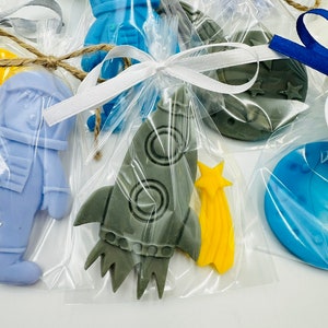 Può includere: Un set di cinque piccoli saponi artigianali colorati a forma di razzo, astronauta, luna e stelle. I saponi sono confezionati in sacchetti di cellophane trasparenti con nastri bianchi e blu.