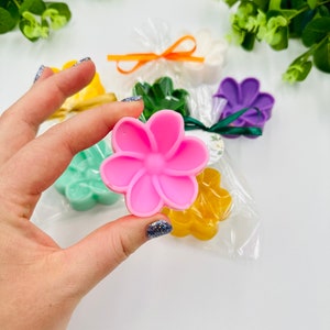 Könnte beinhalten: Sortiment kleiner blütenförmiger Seifen in verschiedenen Farben, darunter rosa, gelb, grün und lila. Die Seifen sind in durchsichtigen Cellophanbeuteln mit Bändern verpackt.