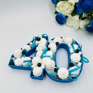 May include: A blue number "40" shaped box filled with white meringue cookies, blueberries, and blue ribbon. A bouquet of white and blue flowers is in the background. The box is a festive decoration.