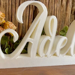 May include: White wooden sign with the name "Adele" in a cursive font. The sign is on a wooden surface with yellow flowers in the background.