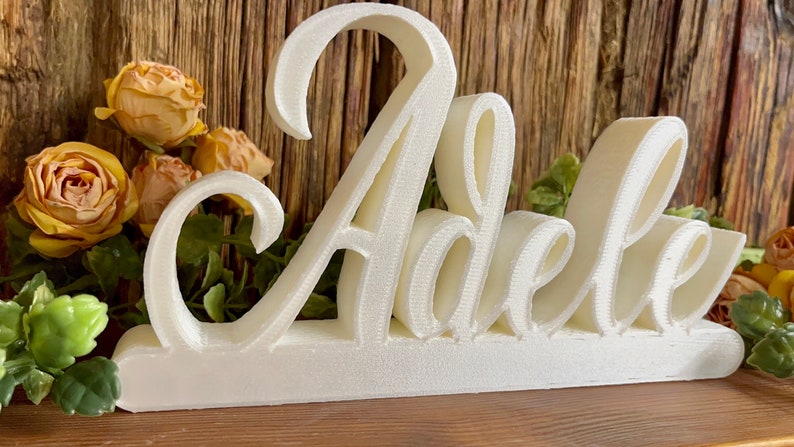 May include: White wooden sign with the name "Adele" in a cursive font. The sign is on a wooden surface with yellow flowers in the background.
