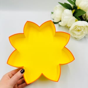May include: A yellow paper flower-shaped box with an orange border. The box is held in a hand.