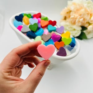 Peut inclure: Un savon rose en forme de cœur tenu dans une main, avec une baignoire blanche remplie de savons colorés en forme de cœur en arrière-plan. Les savons sont de différentes couleurs, dont le rouge, le bleu, le vert et le violet. Un bouquet de fleurs est en arrière-plan.