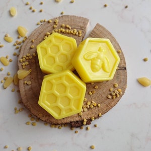 May include: Three yellow, hexagonal soaps with honeycomb and bee designs rest on a wooden slice, surrounded by pollen. The soaps are a bright, sunny yellow color, and the wood has a natural, rustic appearance.