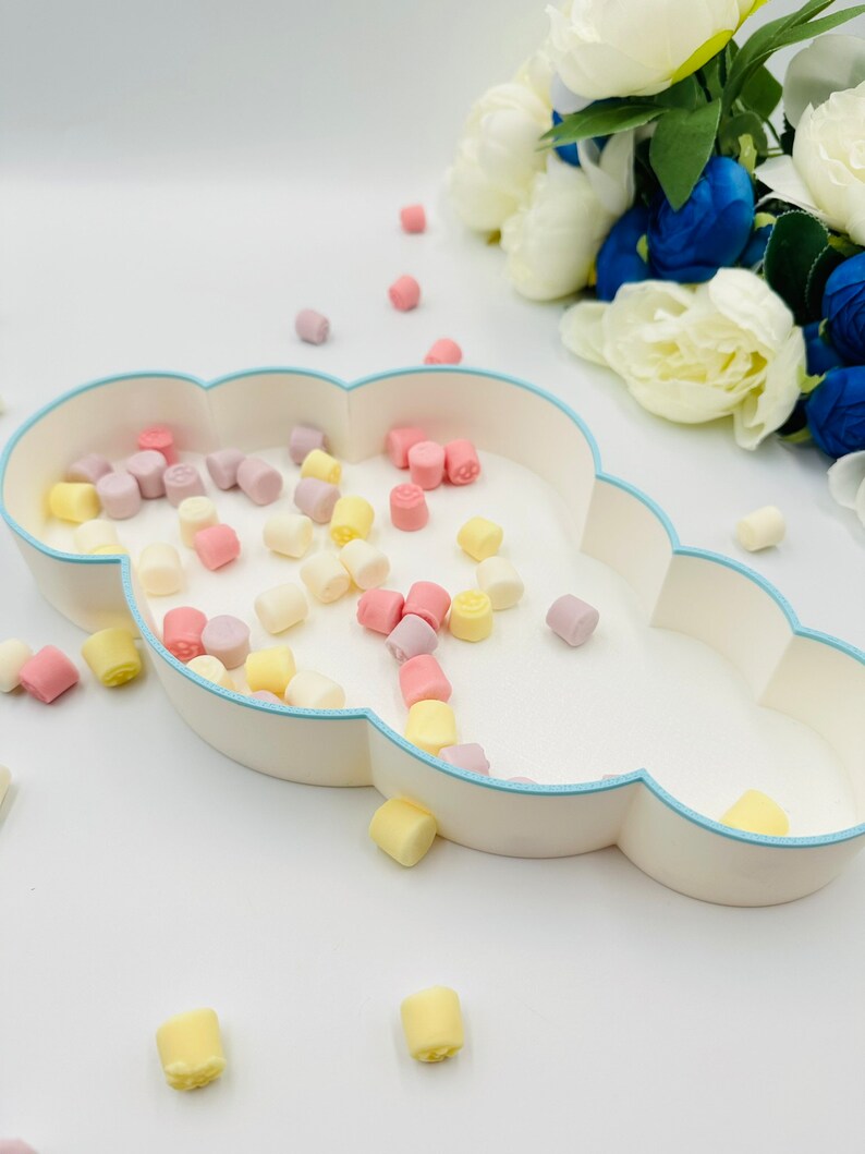 May include: A white cloud-shaped dish with a light blue rim filled with pastel-colored candy. The candy is scattered around the dish on a white surface.