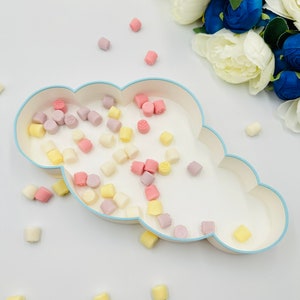 May include: A white cloud-shaped dish with a light blue rim filled with pastel-colored candy. The candy is in the shape of small squares and is scattered around the dish. There are also some candies scattered on the white surface around the dish.