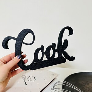 May include: Black wooden sign with the word "Cook" in a cursive font. The sign is held in a hand and is in front of a cookbook and a whisk.