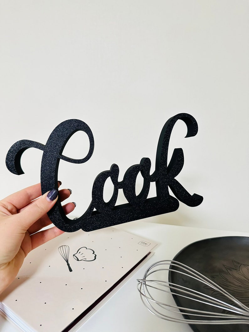 May include: Black wooden sign with the word "Cook" in a cursive font. The sign is held in a hand and is in front of a cookbook and a whisk.