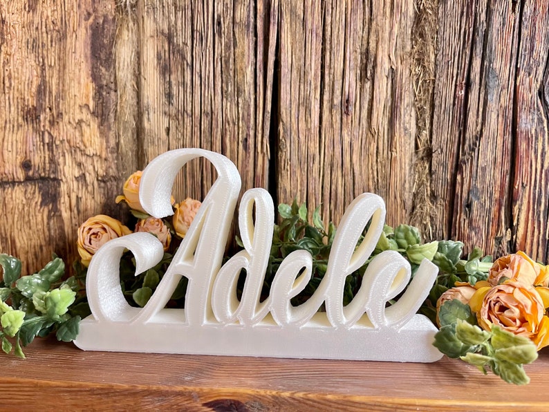 May include: White 3D name sign spelling "Adele" in a cursive font. The sign is on a wooden surface with yellow flowers and green leaves.