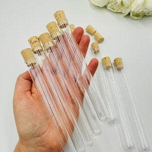 May include: Clear glass test tubes with cork stoppers. The test tubes are held in a hand, with several more test tubes and corks scattered on a white surface. White flowers are in the background.