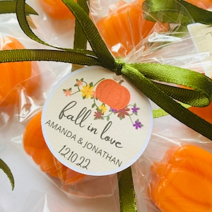May include: Fall wedding favor bags with orange pumpkin-shaped candies, tied with green ribbon and a round tag that says "fall in love" with the couple's names and wedding date, "Amanda & Jonathan 12.10.22".