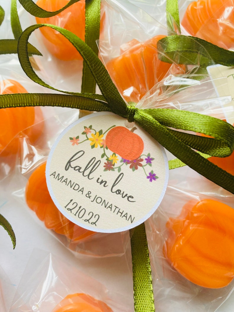 May include: Fall wedding favor bags with orange pumpkin-shaped candies, tied with green ribbon and a round tag that says "fall in love" with the couple's names and wedding date, "Amanda & Jonathan 12.10.22".