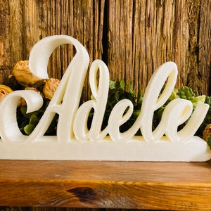May include: White wooden sign with the name "Adele" written in a cursive font.