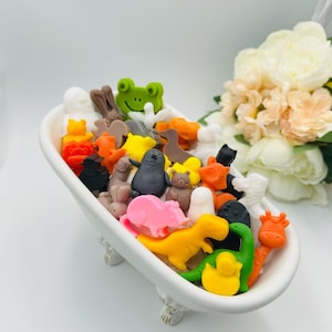 Peut inclure: Une baignoire en céramique blanche remplie de savons colorés en forme d'animaux. Les savons comprennent une grenouille verte, un cochon rose, un dinosaure jaune et un ours brun. La baignoire est sur une surface blanche avec un bouquet de fleurs en arrière-plan.