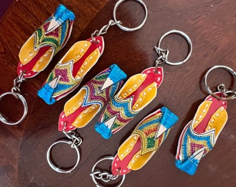 May include: Six colorful keychains shaped like traditional Indian slippers. Each keychain features intricate embroidery in shades of red, yellow, blue, and green. The keychains are attached to silver key rings.