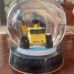 May include: A clear glass snow globe with a yellow and black WALL-E robot inside. The robot has a black visor and is holding a small object in its hand.