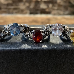 May include: Four silver rings with oval-shaped gemstones. The rings feature a delicate, intricate design with black accents. The gemstones are a deep purple, light blue, red, and yellow.