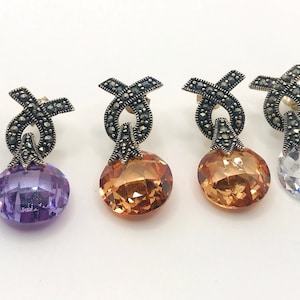 May include: A set of six silver earrings with a black marcasite design and a round crystal stone. The stones are in three colors: purple, orange, and clear.