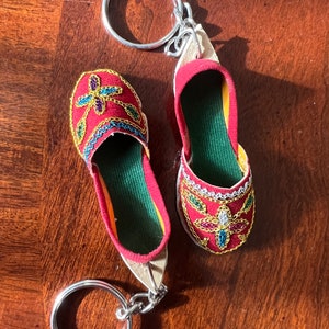 Pode incluir: Um chaveiro de sapato vermelho e dourado bordado com um anel de chave prateado. O sapato tem um forro de tecido verde e enfeites coloridos.