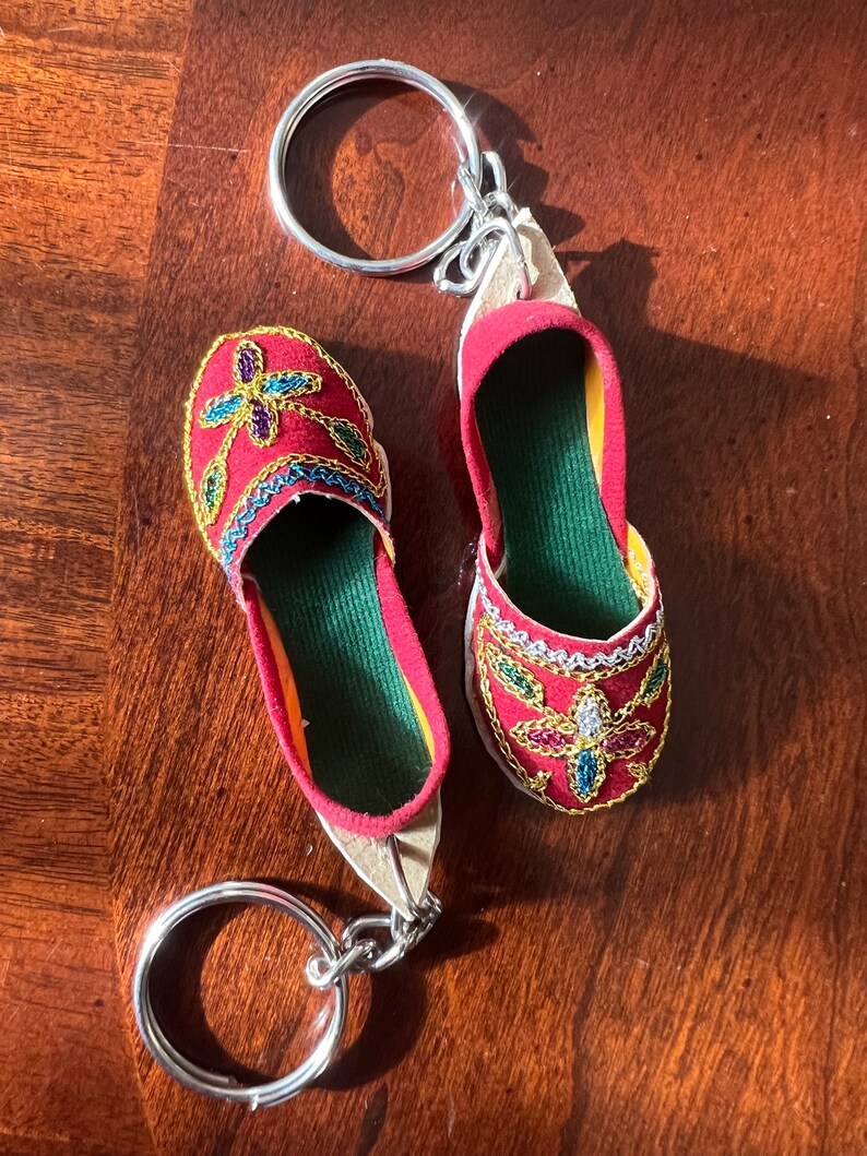 Pode incluir: Um chaveiro de sapato vermelho e dourado bordado com um anel de chave prateado. O sapato tem um forro de tecido verde e enfeites coloridos.
