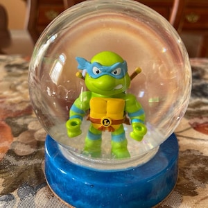 May include: A clear plastic snow globe with a green Teenage Mutant Ninja Turtle figurine inside. The turtle is wearing a blue bandana and holding a katana sword. The snow globe has a blue base.