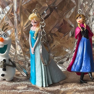 May include: Three Frozen character keychains: Elsa in a light blue dress, Anna in a blue dress with a pink cape, and Olaf the snowman.