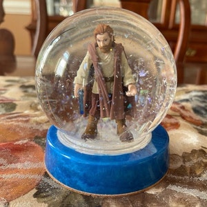 May include: A snow globe featuring a plastic figurine of a Jedi Knight with a brown robe and a lightsaber. The snow globe has a blue base and is filled with white glitter.