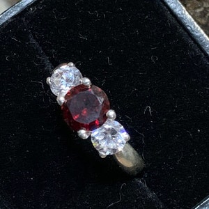 May include: A silver ring with a large red gemstone surrounded by two smaller clear gemstones.