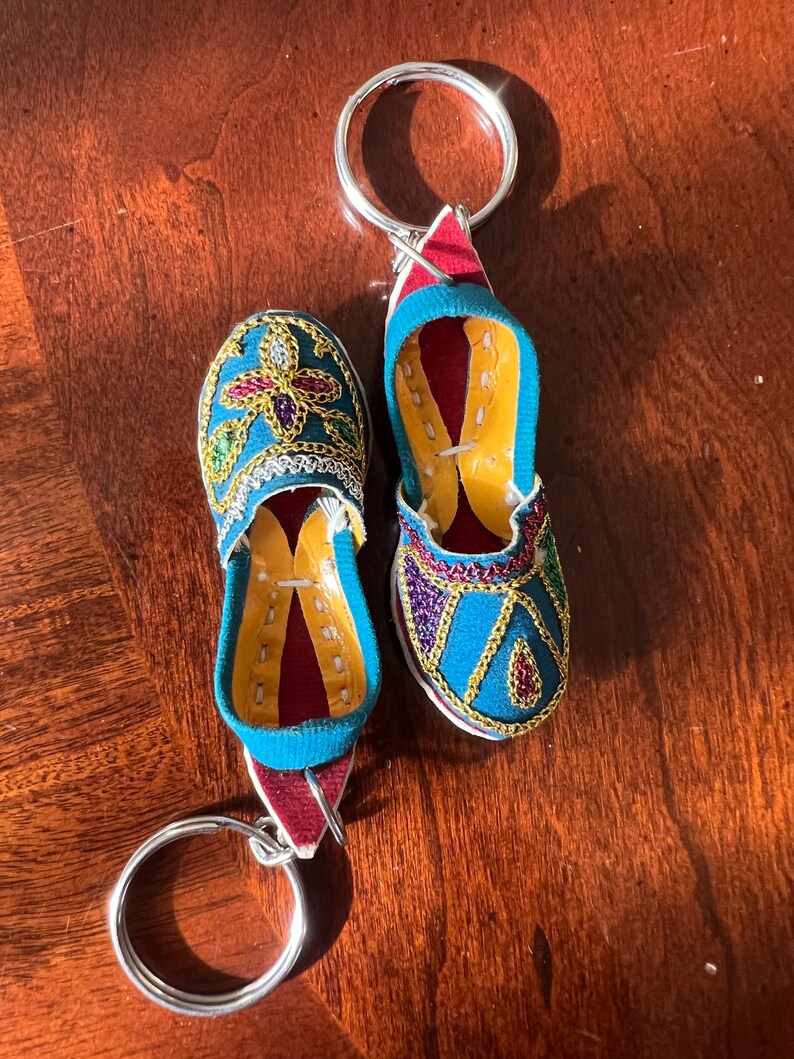 Pode incluir: Um par de chaveiros de pantuflas bordadas em azul e dourado com detalhes vermelhos. Os pingentes s&atilde;o presos a um chaveiro prateado.