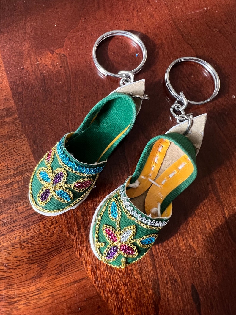 Pode incluir: Um par de chaveiros de pantufla de tecido verde com enfeites de contas coloridas. Os chaveiros est&atilde;o presos a argolas de chave de prata.