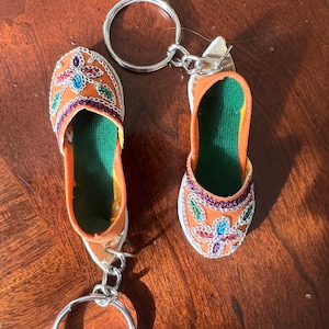 Pode incluir: Um par de chaveiros em forma de chinelo bordado de cor laranja com forro verde e adornos prateados. Os chaveiros est&atilde;o presos a uma corrente e um anel prateados.