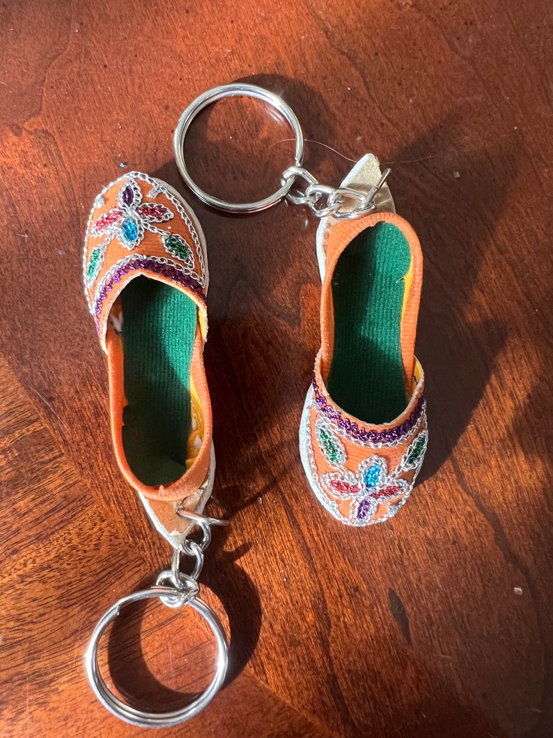 Pode incluir: Um par de chaveiros em forma de chinelo bordado de cor laranja com forro verde e adornos prateados. Os chaveiros est&atilde;o presos a uma corrente e um anel prateados.