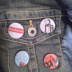 May include: A denim jacket with four button pins. The pins are round and feature various designs and messages. One pin says "Decolonize", another says "Indigenous Peoples for Black Lives", and another features a woman with long dark hair. The fourth pin is a silver pin with the word "Chico's" on it.
